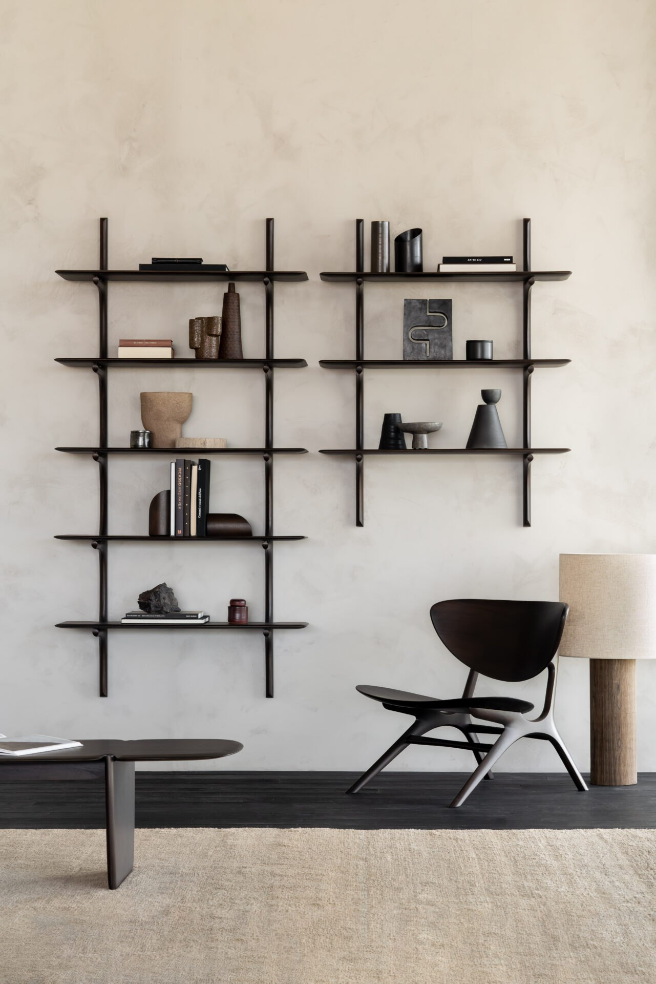 Zwei dunkle PI Wandregale mit Büchern und Keramik über einem Lounge-Sessel und einem niedrigen Tisch.