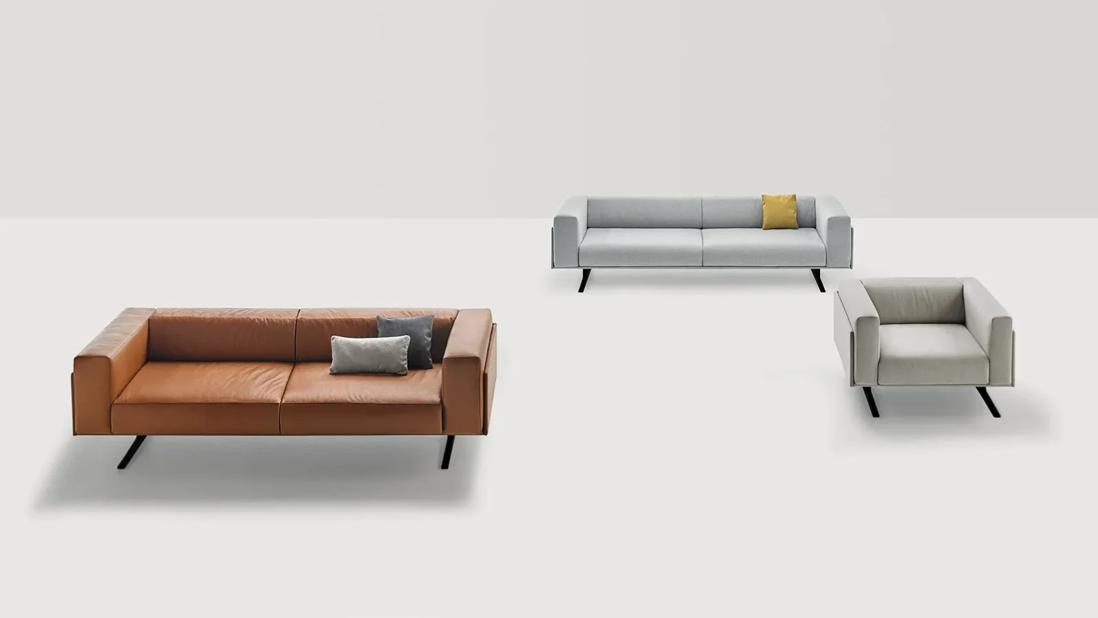 Zwei Marcus Sofas in Braun und Hellgrau sowie ein heller Sessel mit schräg gestellten dunklen Füßen.