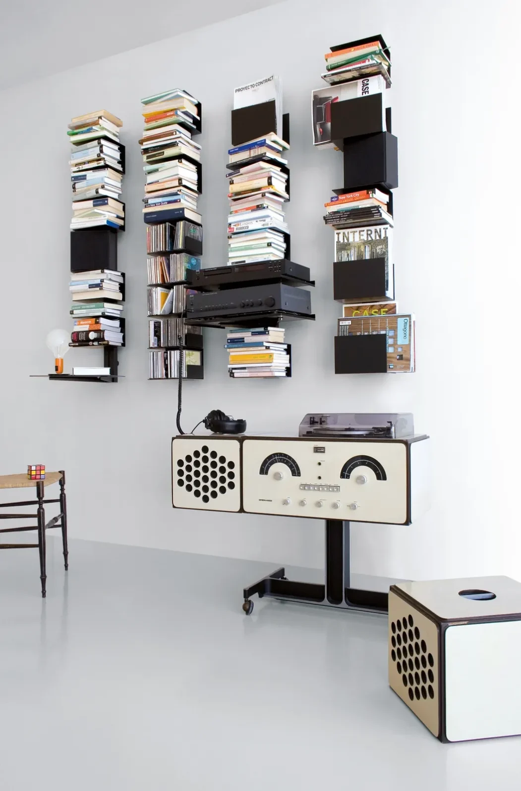 Extendo Wandregal mit schmalen senkrechten Elementen, gestapelten Büchern und Ablagen für Hi-Fi-Geräte.