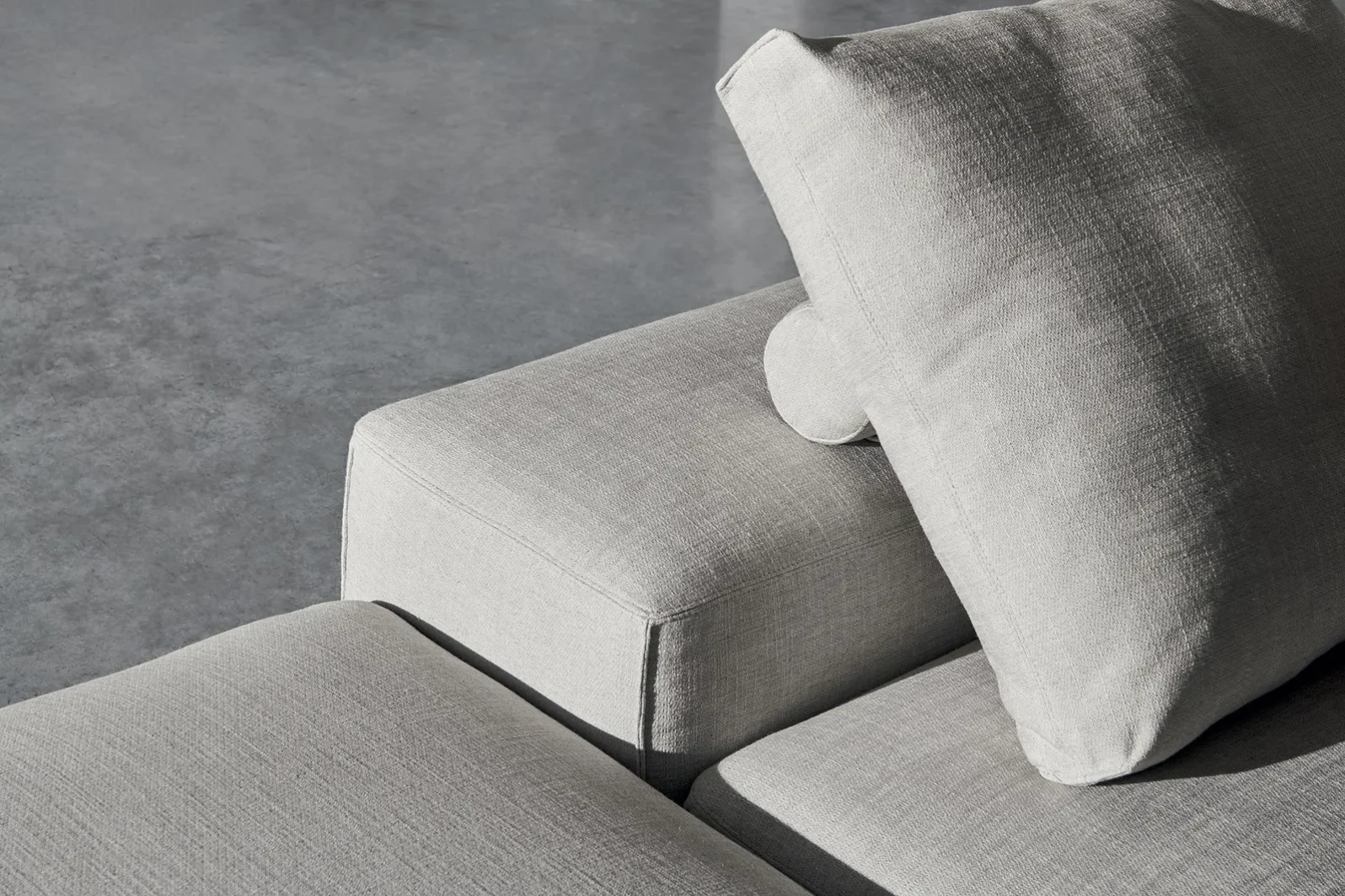 Nahaufnahme des Twils T-Pad Sofas mit grauem Stoffbezug, lockerem Rückenkissen und sichtbarer Naht.